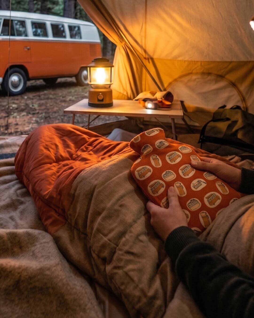 A person inside a tent holding a patterned hot water bottle, with a view of a vintage orange camper van and a glowing lantern outside.