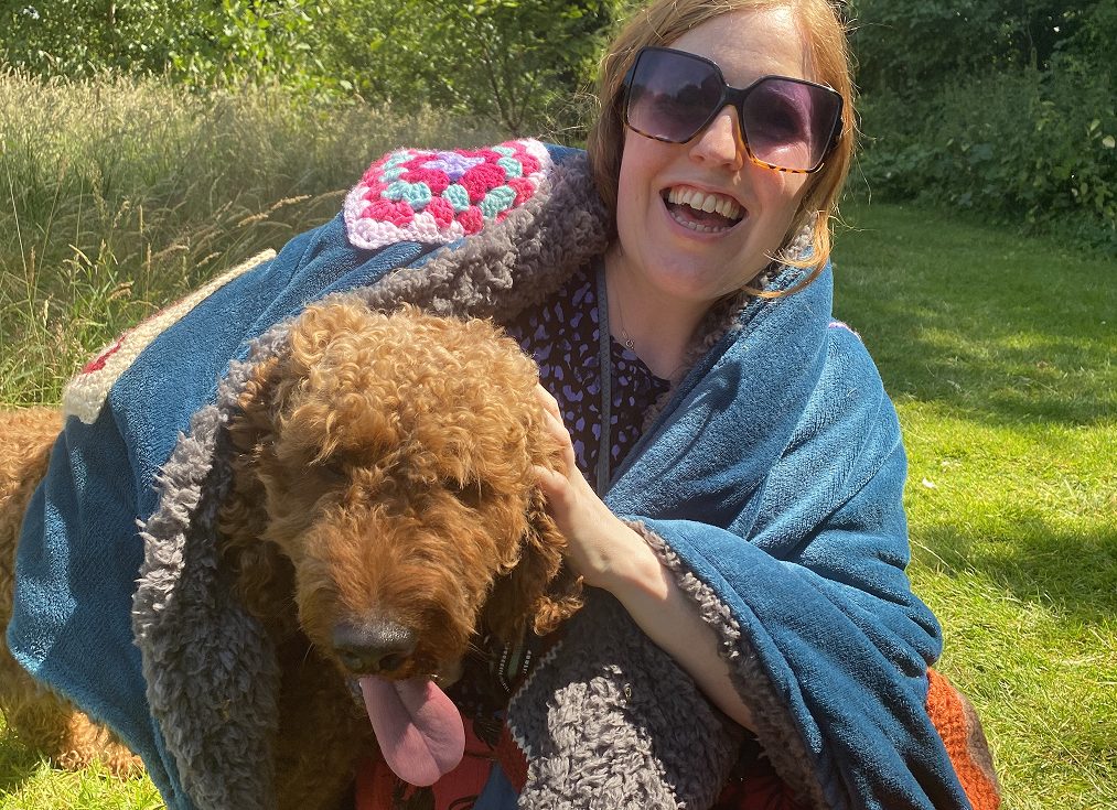 Collette Costello laughing while sitting outdoors in a field, wrapped in a blue crochet-patchwork blanket and hugging a friendly, curly-haired doodle dog.