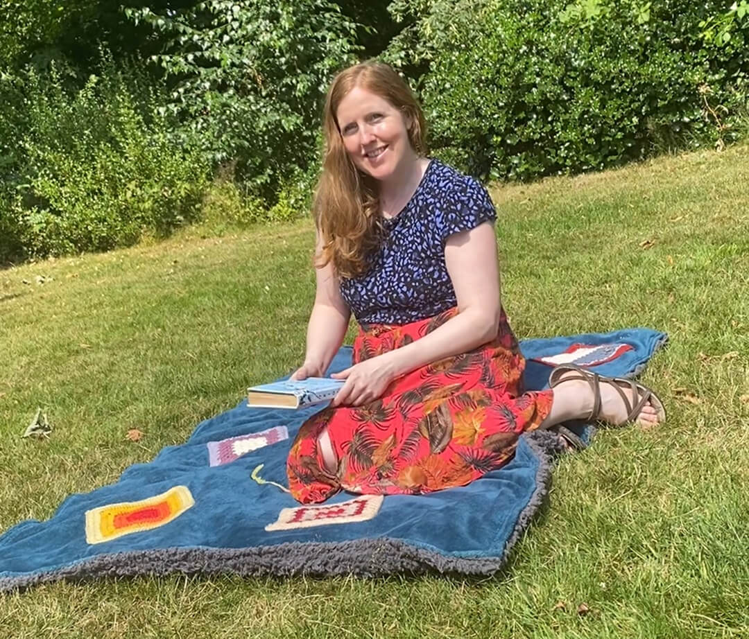 A smiling Collette Costello with ginger hair sits on a plush blue blanket on a sunny green lawn, holding a book during a relaxing outdoor break.