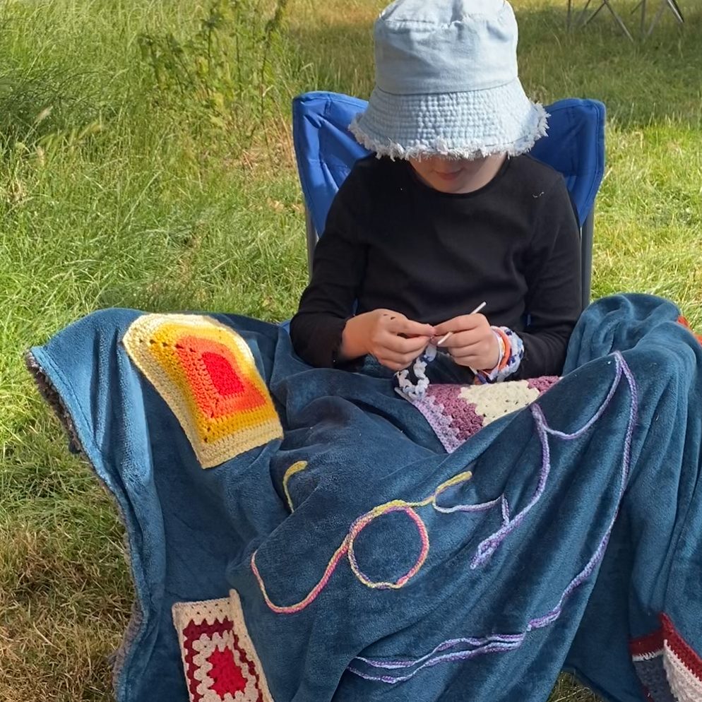 A person sitting in a blue folding chair outdoors on the grass, wearing a light blue bucket hat and a black long-sleeve shirt while crocheting with a hook and yarn. They are wrapped in a cosy blue blanket decorated with colourful granny squares and the word "cosy" stitched across it.