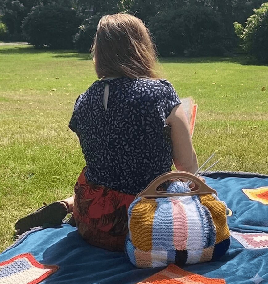 A woman sitting on a picnic blanket in a grassy field next to a handcrafted knit tote bag with wooden handles.