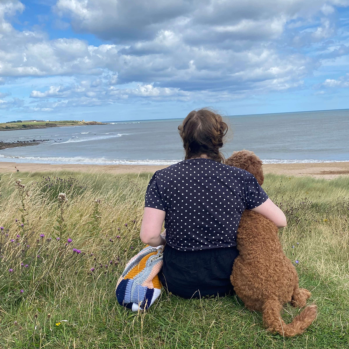 A woman sitting on a grassy dune overlooking the ocean with her arm around a brown curly-coated dog.